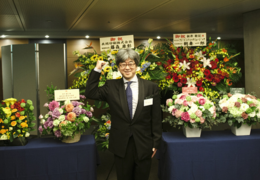 会場に届いた花々の前でポーズをとる新井敏記さん